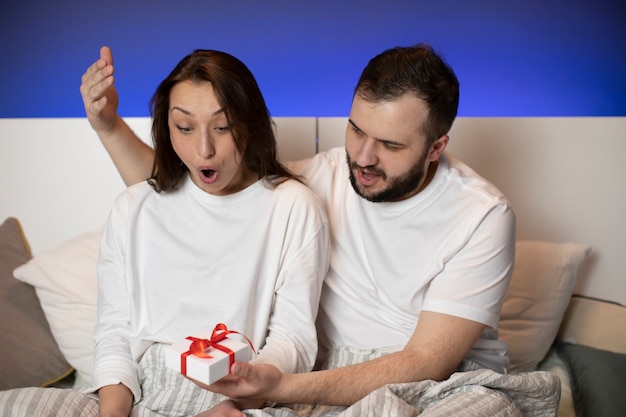 Jeune couple d'amoureux est assis dans son lit, son petit ami présente une boîte-cadeau à une fille surprise
