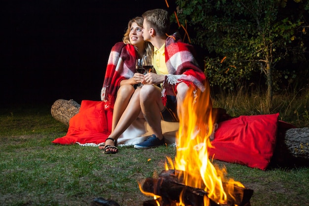 Jeune couple d'amoureux heureux assis sur une bûche près du feu recouvert de plaid tenant du vin, buvant et profitant de leur nuit romantique. il l'embrasse et sourit.