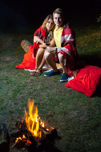 Jeune couple d'amoureux heureux assis sur une bûche près du feu recouvert de plaid tenant du vin, buvant et profitant de leur nuit romantique. regardant la caméra et souriant.