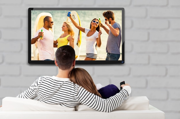 Jeune couple d'amoureux heureux assis sur un canapé à l'intérieur et regardant des photos sur une grande télévision.