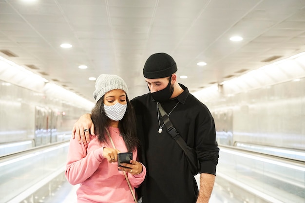 Un jeune couple d'amoureux interracial avec des masques et des chapeaux de laine marchant dans un couloir de métro