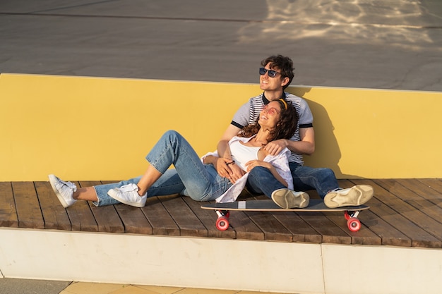 Jeune couple amoureux profiter du coucher du soleil s'asseoir sur un longboard homme et femme à la mode se détendre en plein air