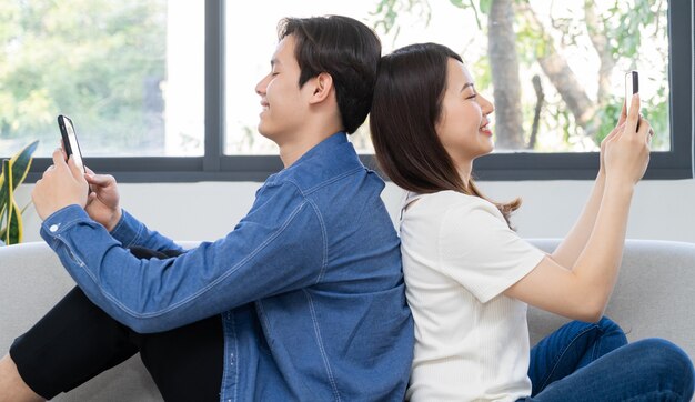 Jeune couple asiatique assis avec le dos l'un sur l'autre et à l'aide du téléphone sur le canapé