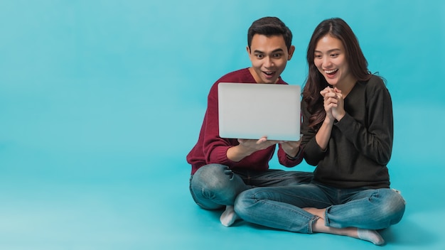 Jeune couple asiatique assis et utilisant un ordinateur portable avec un visage souriant ensemble isolé sur fond bleu. Concept de couple heureux travaillant avec la technologie en ligne.