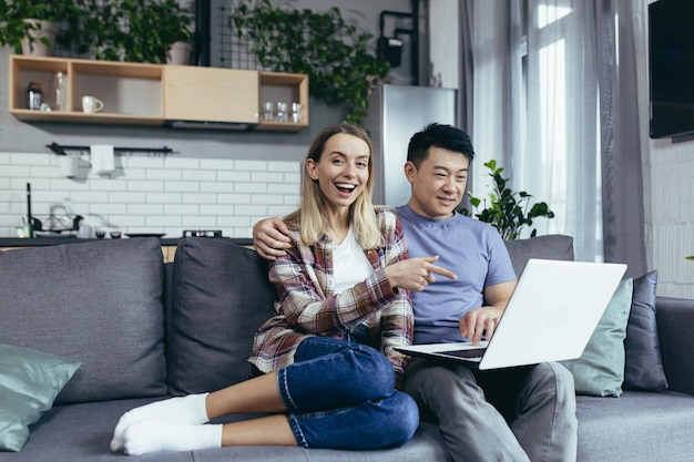 Jeune couple asiatique homme et femme à la maison assis sur un canapé ensemble à l'aide d'un ordinateur portable famille multiraciale heureuse et souriante regardant la caméra