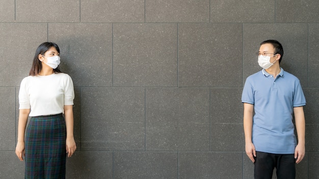 Jeune couple asiatique portant des masques faciaux réunis et debout contre le mur en plein air pour une distance sociale pour le risque d'infection et la prévention des maladies COVID-19.