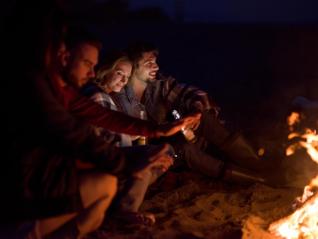 Jeune couple assis avec des amis autour d'un feu de camp sur la plage la nuit en buvant de la bière