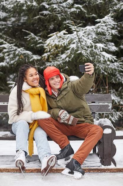 Jeune couple assis sur un banc et faisant portrait de selfie sur téléphone mobile après le patinage dans le parc d'hiver