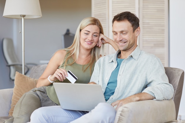 Jeune couple assis ensemble sur un canapé à l'aide d'un ordinateur portable pour payer en ligne avec carte de crédit