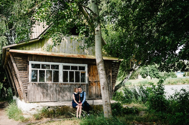 Un jeune couple assis et étreignant tient un livre dans ses mains près d'une vieille maison en bois avec des fenêtres dans la forêt entre les arbres