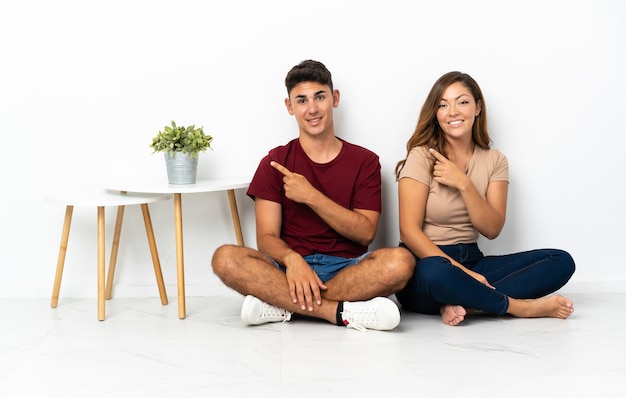 Jeune couple assis sur le sol blanc pointant vers le côté pour présenter un produit