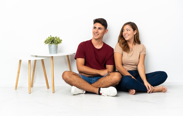 Photo jeune couple assis sur le sol blanc regardant sur le côté et souriant