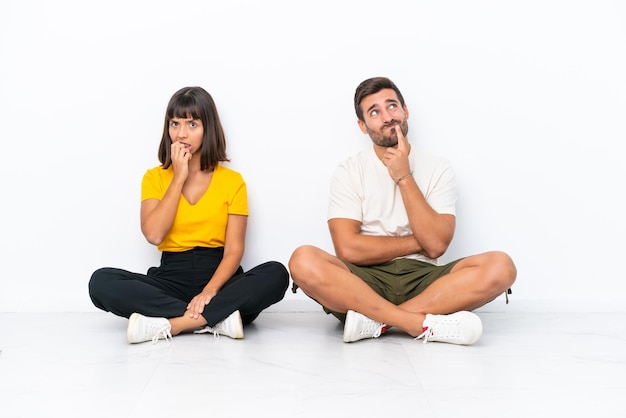 Jeune couple assis sur le sol isolé sur fond blanc ayant des doutes en levant les yeux