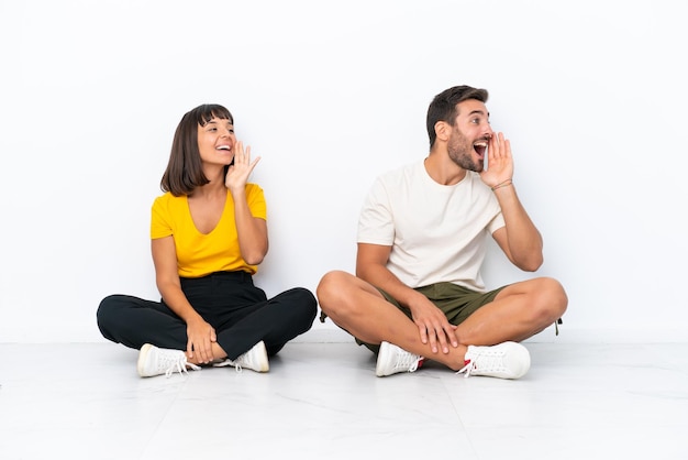 Jeune couple assis sur le sol isolé sur fond blanc criant avec la bouche grande ouverte sur le côté