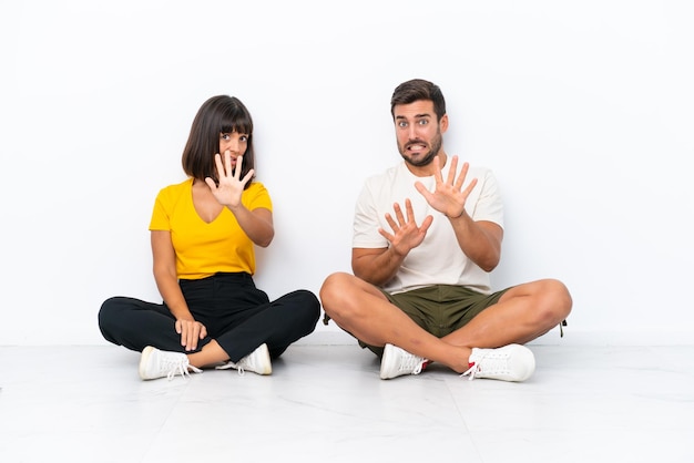 Jeune couple assis sur le sol isolé sur fond blanc est un peu nerveux et effrayé en étirant les mains vers l'avant