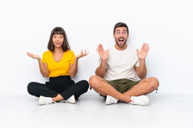 Jeune couple assis sur le sol isolé sur fond blanc avec une expression faciale surprise et choquée