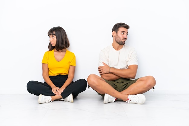 Jeune couple assis sur le sol isolé sur fond blanc avec une expression de visage confus tout en mordant la lèvre