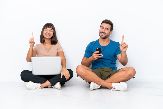 Jeune couple assis sur le sol tenant pc et téléphone portable isolé sur fond blanc montrant et levant un doigt