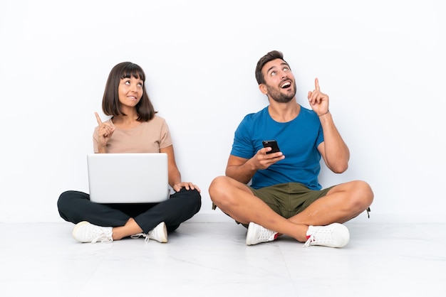 Jeune couple assis sur le sol tenant pc et téléphone portable isolé sur fond blanc pointant avec l'index une excellente idée