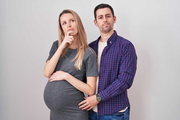 Jeune couple attend un bébé debout sur fond blanc à la confiance à la caméra en souriant avec les bras croisés et la main levée sur le menton pensant positif