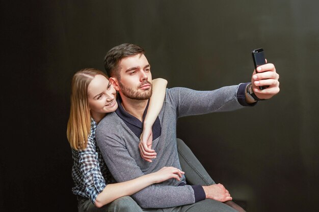 Photo jeune couple de bel homme barbu et femme blonde assise sur fond noir et faisant un drôle de selfie. prise de vue en studio