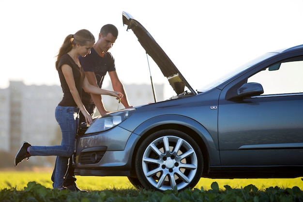 Jeune couple, bel homme et jolie femme à la voiture avec capot sauté, vérification du niveau d'huile dans le moteur. Concept de transport, de problèmes de véhicules et de pannes.