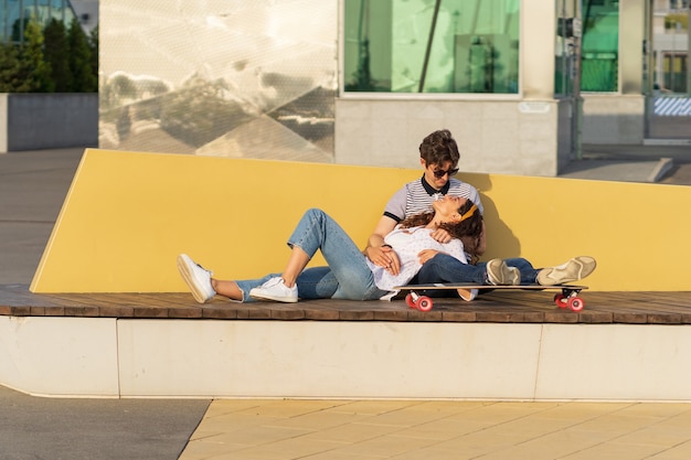 Jeune couple branché heureux câlins parlant s'asseoir sur longboard profiter de l'été en plein air dans le parc urbain de la ville