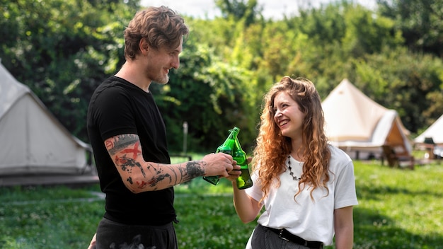 Jeune couple buvant de la bière, souriant, tintant des bouteilles. Verdure autour. Glamping