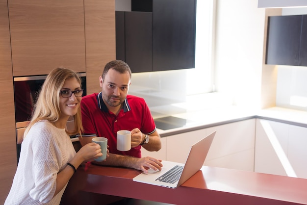 Jeune couple buvant du café et utilisant un ordinateur portable dans une maison de luxe ensemble, regardant l'écran, souriant.