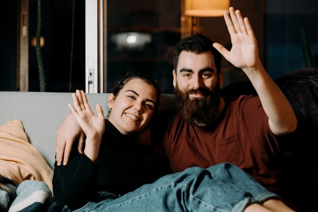 Un jeune couple sur le canapé saluant la caméra et souriant pendant la pandémie