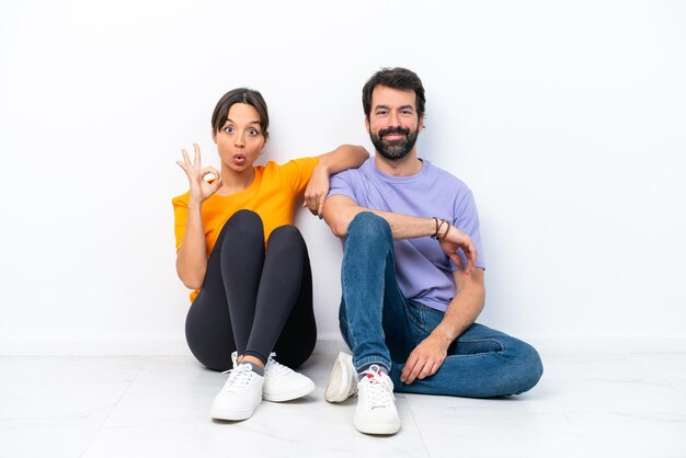 Jeune couple caucasien assis sur le sol isolé sur fond blanc montrant un signe ok avec les doigts