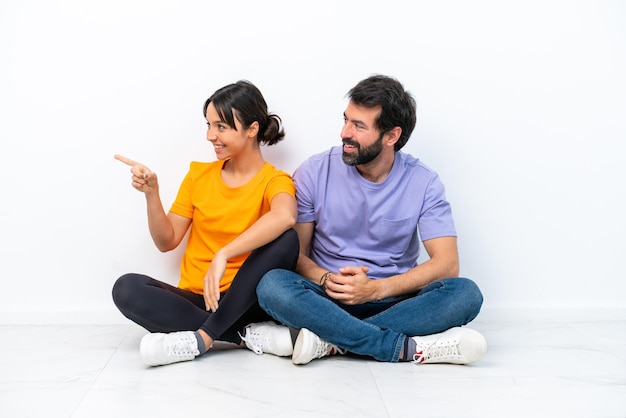 Jeune couple caucasien assis sur le sol isolé sur fond blanc pointant vers le côté pour présenter un produit