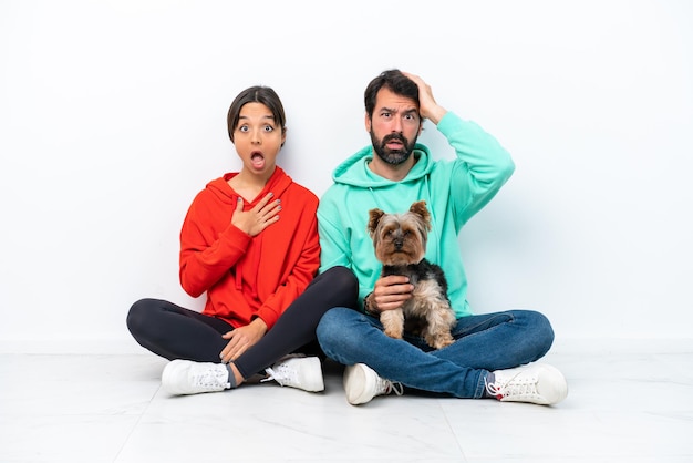 Jeune couple caucasien assis sur le sol avec leur animal de compagnie isolé sur fond blanc avec une expression faciale surprise