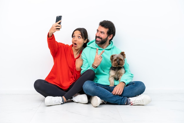 Jeune couple caucasien assis sur le sol avec leur animal de compagnie isolé sur fond blanc faisant un selfie avec le mobile