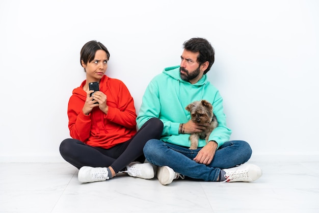 Jeune couple caucasien assis sur le sol avec leur animal de compagnie isolé sur fond blanc lisant un message avec le mobile