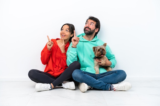 Jeune couple caucasien assis sur le sol avec leur animal de compagnie isolé sur fond blanc pointant vers le haut avec l'index