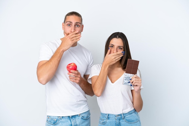 Jeune couple caucasien avec chocolat et pomme isolé sur fond blanc couvrant la bouche avec les mains pour dire quelque chose d'inapproprié