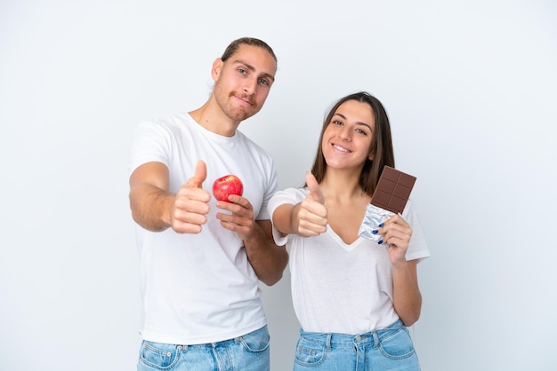 Jeune couple caucasien avec chocolat et pomme isolé sur fond blanc donnant un coup de pouce parce que quelque chose de bien s'est produit