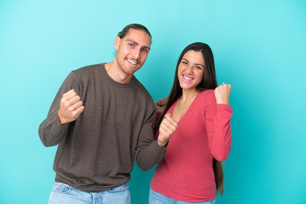 Jeune couple caucasien isolé sur fond bleu célébrant une victoire en position de vainqueur