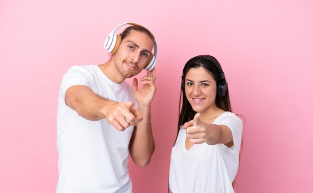 Jeune couple caucasien isolé sur fond rose écoutant de la musique et pointant vers l'avant