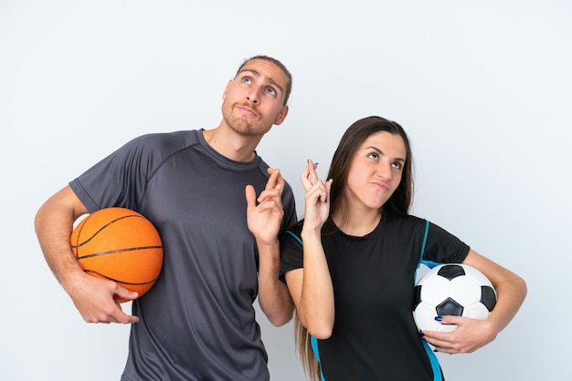 Jeune couple caucasien jouant au basket et au football isolé sur fond blanc avec les doigts croisés et souhaitant le meilleur