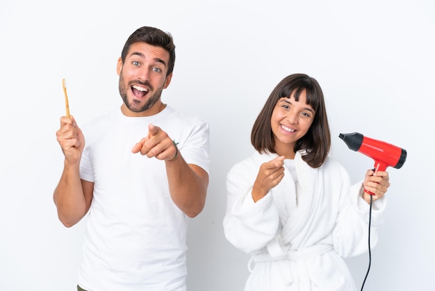 Jeune couple caucasien tenant un sèche-cheveux et une brosse à dents isolé sur fond blanc pointe le doigt vers vous avec une expression confiante