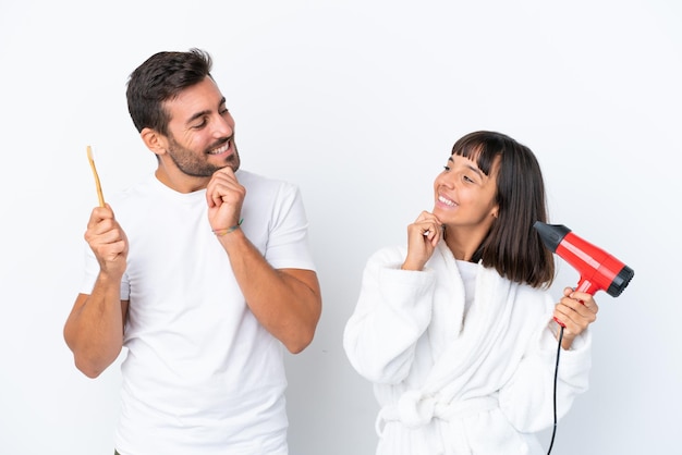 Jeune couple caucasien tenant un sèche-cheveux et une brosse à dents isolés sur fond blanc regardant les uns les autres