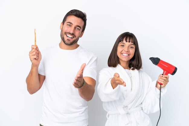 Jeune couple caucasien tenant un sèche-cheveux et une brosse à dents isolés sur fond blanc se serrant la main pour conclure une bonne affaire