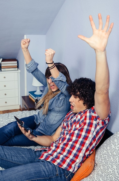 Jeune couple célébrant la victoire en regardant le sport sur la tablette assis sur le lit