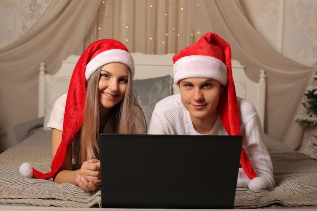 un jeune couple en chapeaux de père noël est allongé sur le lit à la maison avec un ordinateur portable noël