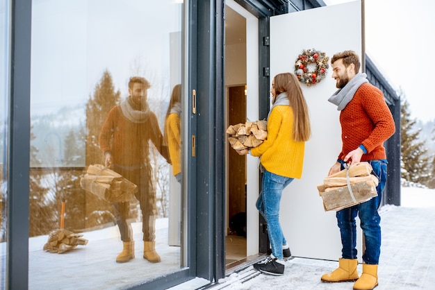 Jeune couple charmant vêtu de chandails colorés entrant dans leur maison moderne avec du bois de chauffage dans les montagnes pendant l'hiver