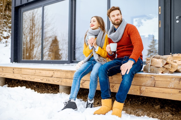 Jeune couple charmant vêtu de pulls colorés profitant de la nature assis avec des boissons chaudes sur la terrasse de la maison moderne dans les montagnes