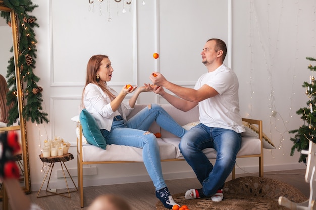 Jeune couple en chemises blanches et jeans s'amusant avec des mandarines sur un canapé contre un sapin de Noël décoré à la maison. Vacances d'hiver au concept de maison confortable..