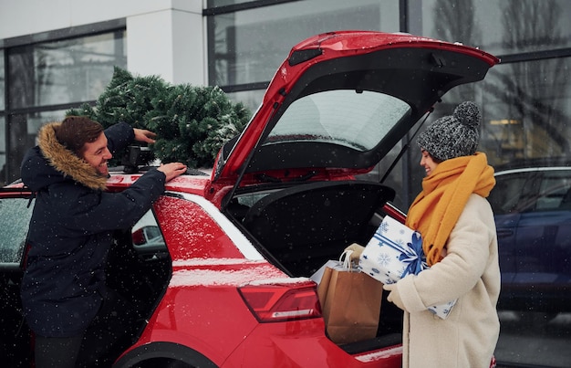 Un jeune couple avec des coffrets cadeaux est près d'une voiture avec un arbre sur le dessus. Ensemble à l'extérieur à l'heure d'hiver.